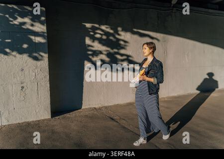 donna che cammina con la baguette alla luce del sole e getta lunghe ombre su una parete di cemento Foto Stock