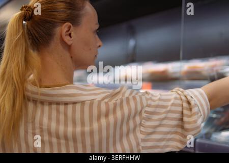 Donna in piedi in una sezione refrigerata del supermercato, in cerca di articoli confezionati, concentrandosi sulla spesa e facendo decisio di acquisto quotidiano Foto Stock