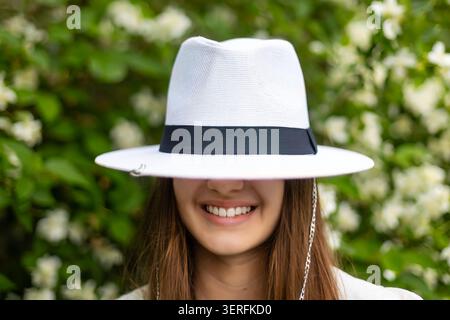 Giovane donna sorridente che indossa un cappello bianco in giardino. La giovane donna sorridente indossa un cappello bianco basso sugli occhi con fiori bianchi in fiore e fogliame verde Foto Stock