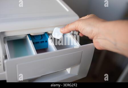 Primo piano della mano di una persona posizionando una cialda di detergente multicolore nell'erogatore a cassetto aperto di una lavatrice bianca Foto Stock