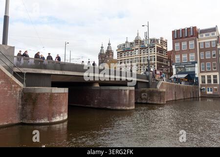 AMSTERDAM, PAESI BASSI - 10 NOVEMBRE 2025: Persone in piedi su un ponte che si affaccia su un canale con edifici storici ad Amsterdam Foto Stock