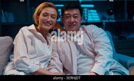 Felice allegra coppia multirazziale guardare la televisione ridendo godendo di trascorrere il tempo insieme in salotto di sera coniugi multietnici uomo asiatico Foto Stock