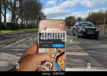 Mano che tiene in mano lo smartphone che mostra l'app Domino per la consegna della pizza con la strada in background Foto Stock