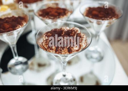 Il dessert al cioccolato si siede in eleganti tazze di vetro. Ogni porzione è ricoperta da un ricco cacao in polvere. I bicchieri poggiano su una superficie bianca pulita. Illuminazione soffusa alta Foto Stock