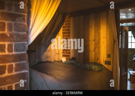 Un comodo letto con cuscini a motivi geometrici è illuminato da una calda luce ambientale, creando un'atmosfera intima in una camera rustica Foto Stock