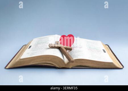 Cuore di carta rossa e croce crocifisso in legno in cima alla bibbia aperta. Isolato su sfondo azzurro. Foto Stock