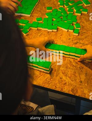 Le mani dei giocatori impilano e spingono file di piastrelle Mahjong con il retro verde verso il basso per costruire muri su un tavolo in truciolato di legno. Foto Stock