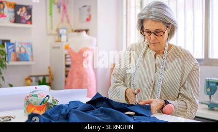 Donna dai capelli grigi che taglia il tessuto in un'officina di cucito Foto Stock