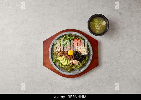 Bibimbap dolsot coreano servito in un tradizionale vaso di pietra su un vassoio di legno con tuorlo d'uovo, verdure assortite, alghe e manzo, accompagnato da una ciotola Foto Stock