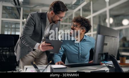 Due uomini d'affari che esaminano le informazioni in tablet digitale e computer ridendo divertente felice parlare uomo caucasico boss che mostra i dati online a. Foto Stock