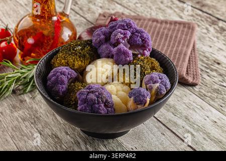 Fiori di cavolfiore e broccoli congelati in una ciotola nera su un tavolo di legno. Verdure viola, bianche e verdi con olio, pomodori e rosmarino nea Foto Stock