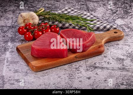 Due filetti di tonno crudo su un tagliere di legno. Gli ingredienti freschi per cucinare includono pomodori ciliegini, aglio e rosmarino. Preparazione sana dei pasti su rustico gre Foto Stock