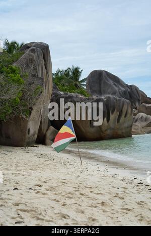 La bandiera nazionale piantata su una spiaggia di sabbia bianca vicino a iconici massi di granito, Anse Source d'Argent, La Digue, Seychelles Foto Stock