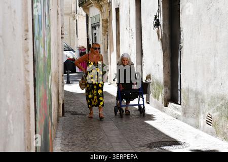 Signore che camminano lungo una strada illuminata dal sole nella città francese di Beaucaire in Provenza, Francia. Una donna sta usando una cornice per camminare per aiutarla a camminare. Foto Stock
