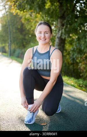 Donna che lega lacci e sorride prima di correre. Donna runner che si prepara sul percorso all'aperto, umore positivo, preparazione, allenamento fitness, stile di vita attivo, concetto di motivazione Foto Stock