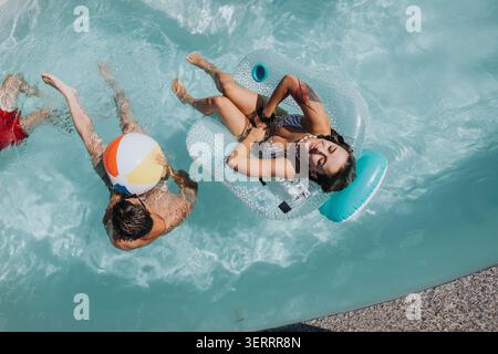 Giovane donna che galleggia su un gonfiabile in piscina mentre un amico gioca con un pallone da spiaggia Foto Stock