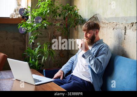 Un lavoratore freelance attento che tiene la tazza accanto al notebook in un accogliente spazio caffetteria, esprime calma nella concentrazione, pausa di lavoro, processo decisionale e concetto di slow living moderno Foto Stock