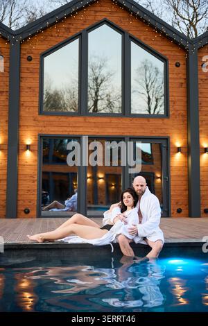 Donna che riposa sulla grembo del partner sulla terrazza a bordo piscina. Coppia che gode di un intimo rifugio spa con luci calde, intimità, relax e concetto di fuga di lusso Foto Stock