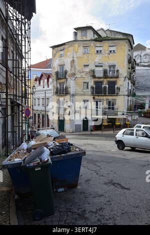 Scena uban con edifici residenziali obsoleti, auto parcheggiate e contenitori di rifiuti straripanti in una strada nel centro storico di Lisbona, Portogallo. Foto Stock