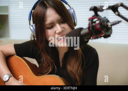Primo piano, una musicista asiatica che pratica la chitarra acustica canta in studio con cuffie e microfono registrazione, emozionale f Foto Stock