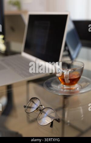 Un paio di occhiali da vista con struttura in metallo poggiano su un tavolo di vetro con riflessi per il tè e il laptop Foto Stock