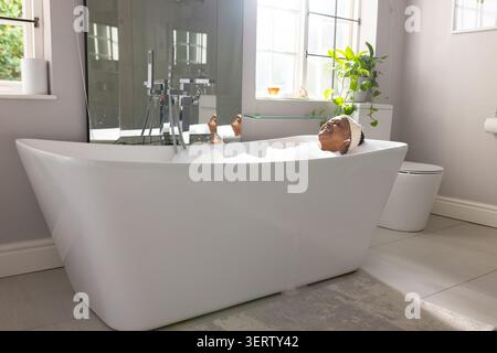 Vasca da bagno indipendente bianca che tiene le bolle vicino alle finestre, stucco cromato riflesso nello specchio Foto Stock
