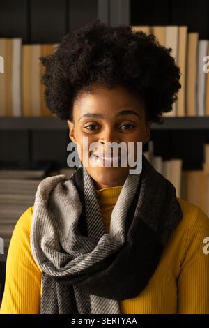 Donna che sorride e indossa dolcevita a coste senape e sciarpa grigia a maglia nello studio domestico con i libri Foto Stock