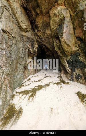 Vista di una persona con uno zaino che cammina attraverso una grande grotta marina con pareti di granito e alghe marine sul pavimento sabbioso della baia di Nanjizal a Saint Just, Eng Foto Stock