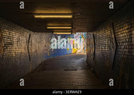 Kaarst, Germania, 10.06.2026 - tunnel sotterraneo pedonale urbano scuro con graffiti sulle pareti in mattoni e plafoniere con illuminazione soffusa che conduce ad un'uscita luminosa. Foto Stock