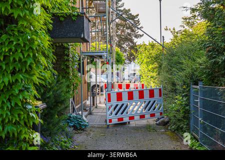 Kaarst, Germania, 10.06.2026 - ponteggi e barriera di sicurezza su una stretta strada residenziale accanto alla facciata di una casa coperta di edera verde Foto Stock
