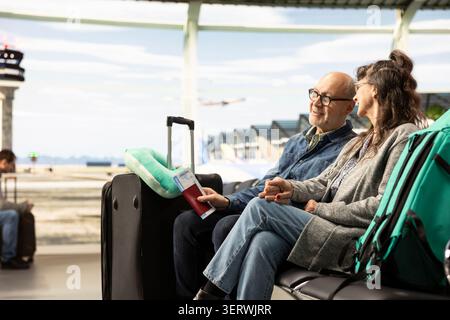 Felice coppia senior seduta insieme nella lounge dell'aeroporto in attesa con i bagagli, sorridendo e chiacchierando mentre controlla le informazioni sulla partenza del volo. I passeggeri anziani abbracciano la libertà di pensionamento. Foto Stock