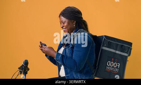 Donna che esplora il telefono cellulare mentre è in bicicletta pronta per il servizio di consegna del cibo, con una giovane donna che abbraccia l'economia frenetica utilizzando lo smartphone per la navigazione. Foto Stock