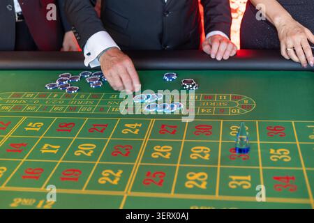 La gente si riunisce intorno a un tavolo da roulette in un casinò. I giocatori piazzano le proprie chip sul layout delle scommesse. L'atmosfera e' vivace con eccitamento mentre si rilassano Foto Stock