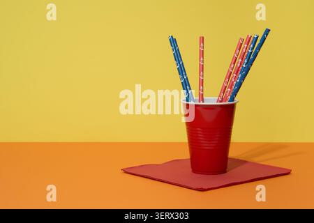 Un bicchiere di plastica rosso, cannucce blu e rosse e un tovagliolo su un tavolo arancione con sfondo giallo Foto Stock