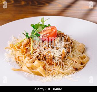 Tagliatelle condite con un ricco sugo di carne alla bolognese, parmigiano grattugiato, una fetta di pomodoro fresco e contorno verde. Presentato su un piatto bianco appoggiato su un tavolo di legno, perfetto per un pasto. Foto Stock