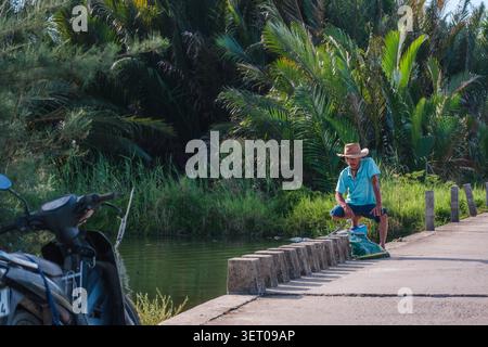 Uomo asiatico in un cappello di paglia appoggiato su un ponte di cemento sopra un fiume Foto Stock