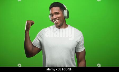 L'uomo nero che indossa le cuffie si flette bicipite destro e sorride verso la fotocamera in uno studio green screen indossando una t shirt bianca; un ritmo sicuro. Foto Stock