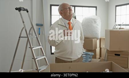 Uomo anziano che tiene la penna al mento tra le scatole di cartone e la scala all'interno di un edificio, indossando occhiali e cravatta arancione; pianificazione nostalgica. Foto Stock