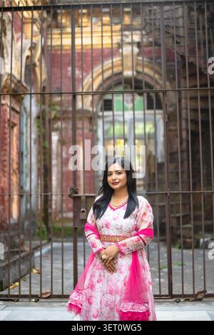 Una donna in un vestito a motivi rosa e bianchi con uno scialle nero si trova davanti a un edificio recintato con finestre ad arco e ferri decorativi. Foto Stock