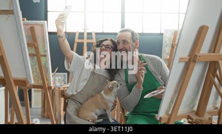 Una donna di mezza età solleva lo smartphone per un selfie in uno studio d'arte con un uomo che dipinge su un cavalletto e un piccolo cane in grembo; gioia di compagnia. Foto Stock
