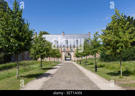 Un sentiero acciottolato alberato conduce al cancello d'ingresso dell'abbazia di Abdij van Park a Lovanio, Belgio, incorniciato da edifici storici in mattoni e prato verde Foto Stock