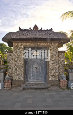 Un ingresso a una villa di lusso realizzata con pareti in pietra corallina rustica, caratterizzato da una doppia porta in legno grigio splendidamente intagliata e da un tetto di paglia a Bali Foto Stock