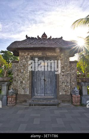 Un ingresso a una villa di lusso realizzata con pareti in pietra corallina rustica, caratterizzato da una doppia porta in legno grigio splendidamente intagliata e da un tetto di paglia a Bali Foto Stock