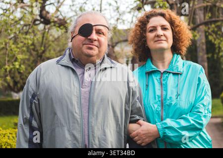 Coppia di mezza età che cammina insieme a un uomo che indossa un cerotto per gli occhi e un compagno di supporto all'aperto Foto Stock