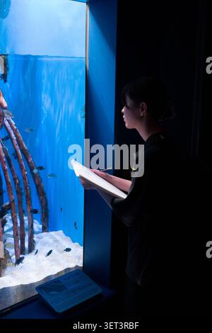 Una giovane donna che disegna con un taccuino mentre guarda una vasca blu in un acquario. Foto Stock