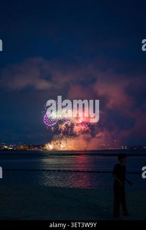 Vivaci fuochi d'artificio che esplodono sull'oceano di notte con luci della città in lontananza e la silhouette di una persona su una spiaggia. Foto Stock