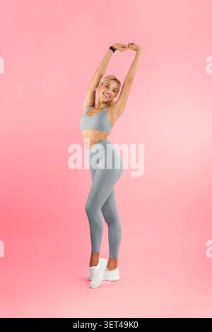 Happy Young Woman che si allunga in Sportswear, outfit grigio, sfondo Pink Studio, fitness Concept Foto Stock