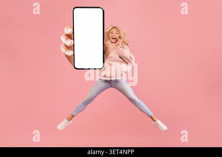 Felice giovane donna che salta con smartphone che visualizza schermo vuoto, sfondo rosa, Mockup Foto Stock