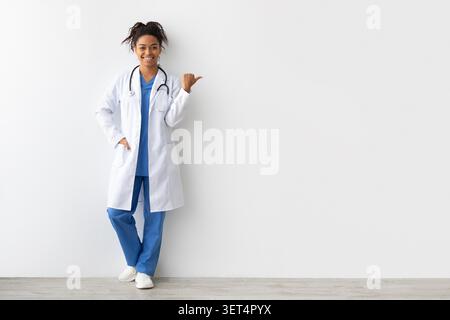 Medico femminile sorridente che indossa camice da laboratorio e scrub, puntando con gesto della mano, concetto Clinic Healthcare Foto Stock