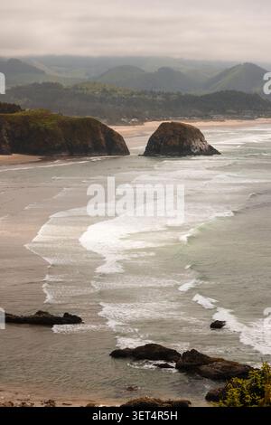 Onde che si infrangono sulla costa sabbiosa dell'Oregon con l'iconica Haystack Rock e colline sotto cieli nuvolosi Foto Stock